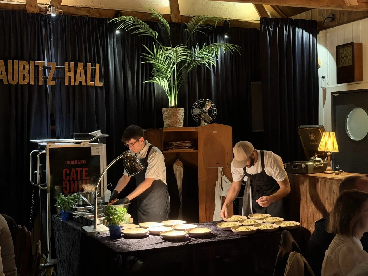 To kokker forbereder mat på en serveringsdisk med flere ferdige retter. Bakgrunn med svart gardin og en stor grønn plante.