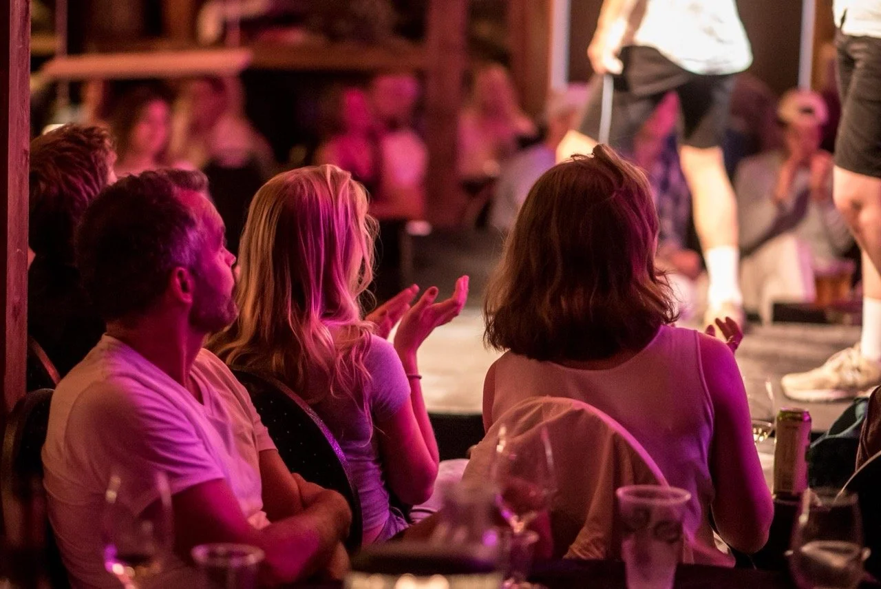 En gruppe mennesker sitter i en mørk, lystig scene, ser på en opptrender på scenen, med rosa og oransje belysning. Noen av dem applauderer.