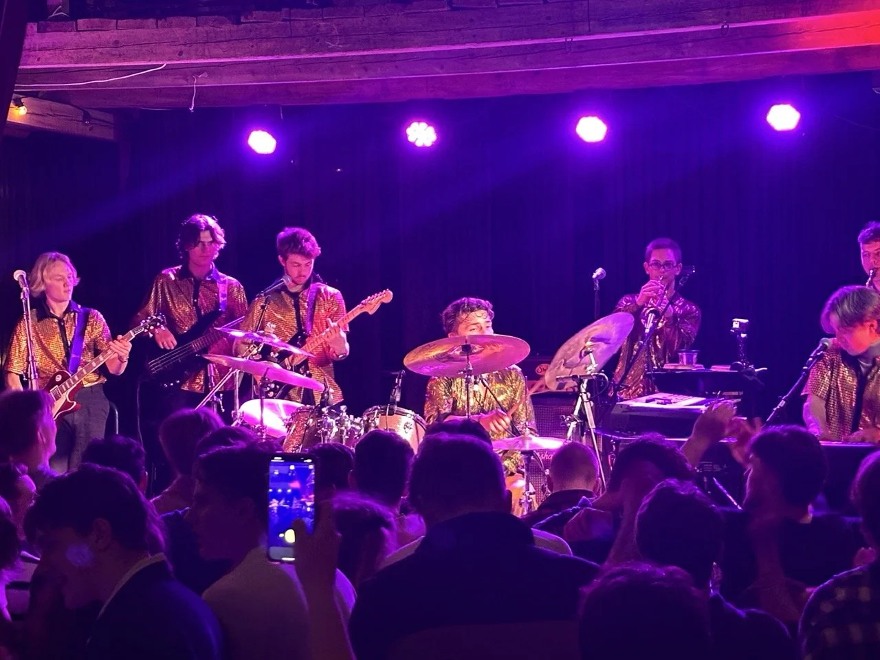 Musikkgruppe som spiller på scenen med publikum foran, i et rom med mørke og rosa lys.