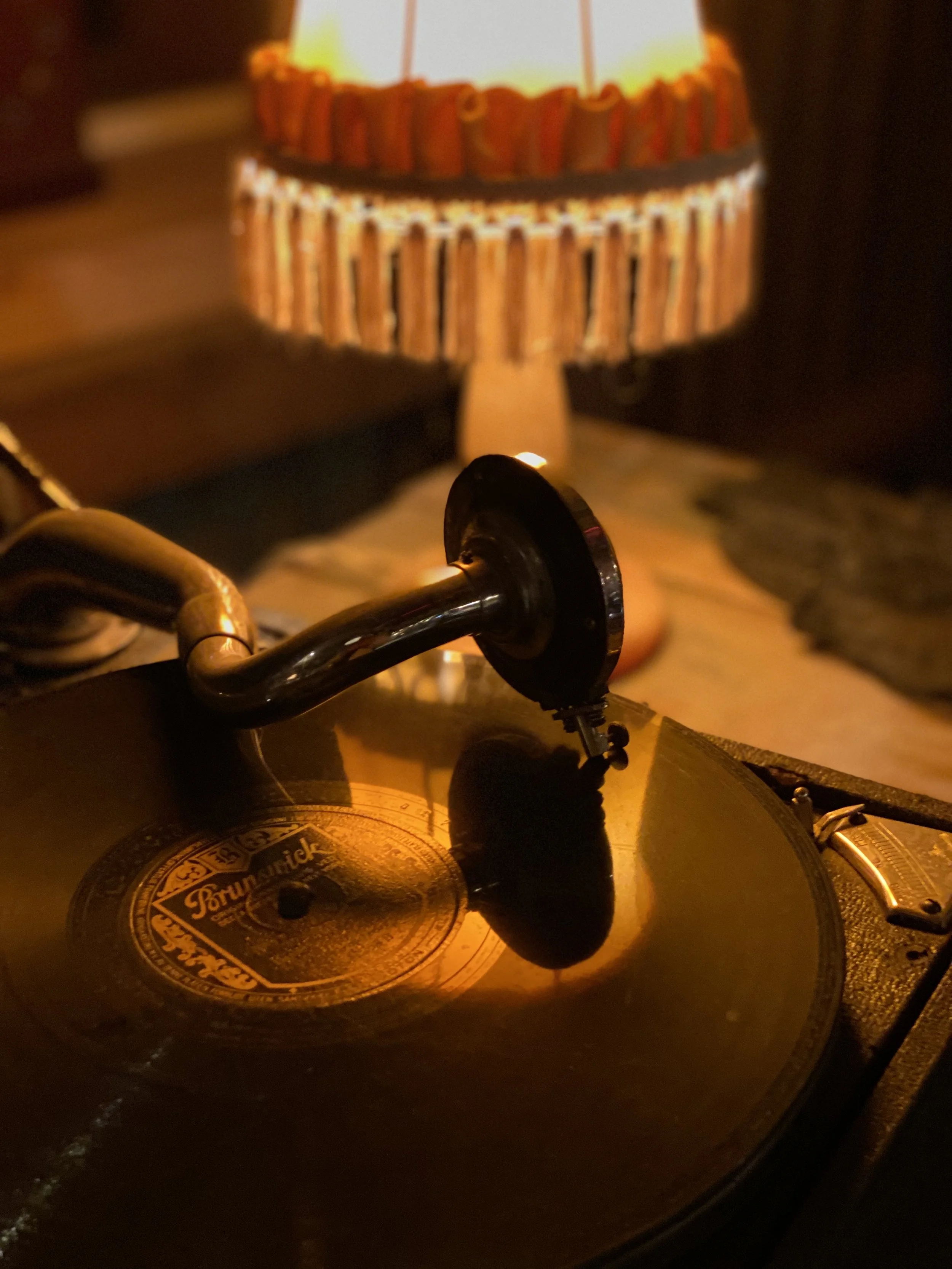 Nærbilde av en plateplate av en grammofon, med en tonearm og nål, plassert på et bord. I bakgrunnen er det en bordlampe med en fysisk skjerm.