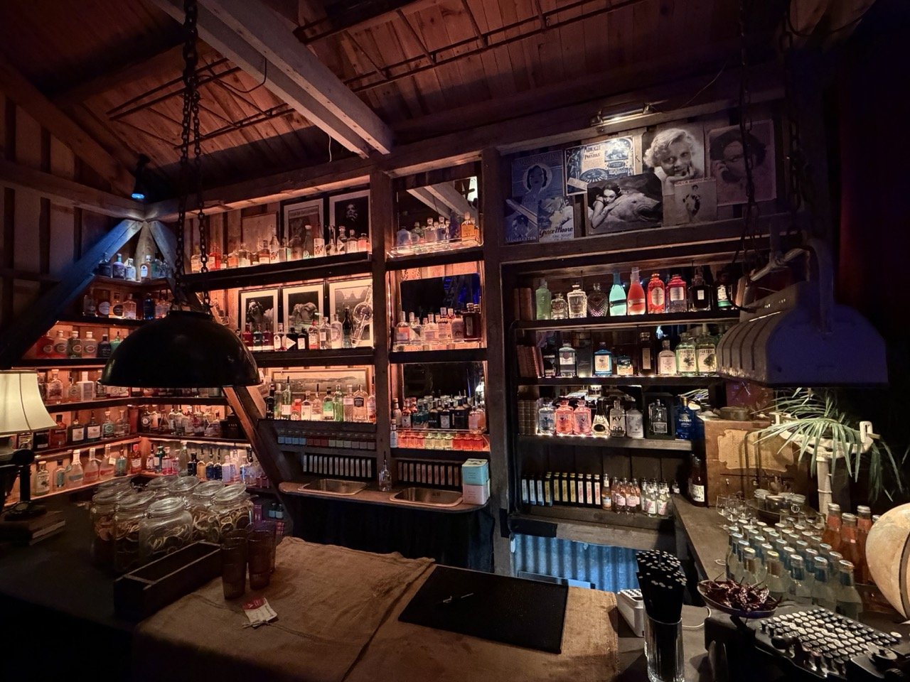 Interiør av et hjemmekoselig bar- eller samlingsrom med mange alkoholflasker på hyller, dekorative bilder på veggene, et lite vaskekar under hyllene, og en samling av glass og pynt på bordet.