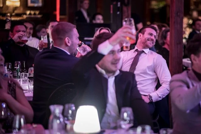 Publikum i et festlig selskap som drikker og smiler under en kveld med underholdning.