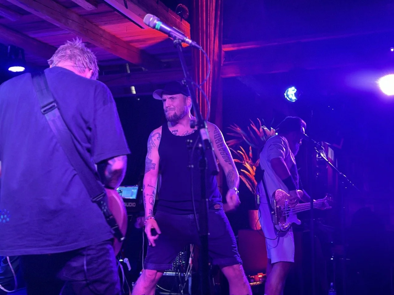 Tre bandmedlemmer som opptrer på scenen i en bar eller klubb med fargerik belysning.