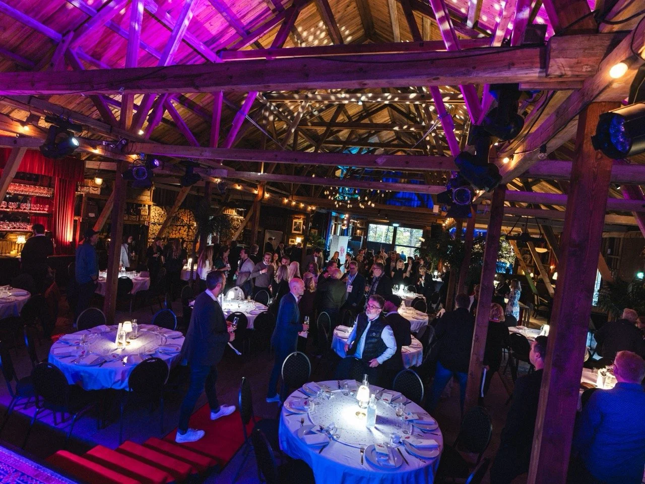 Festlokale med rundt bord dekket med hvite duker og spiselige sette, folk som mingler og prater, dekorasjoner med lystelys og trestolper, et barområde i bakgrunnen, fargerik belysning på taket.
