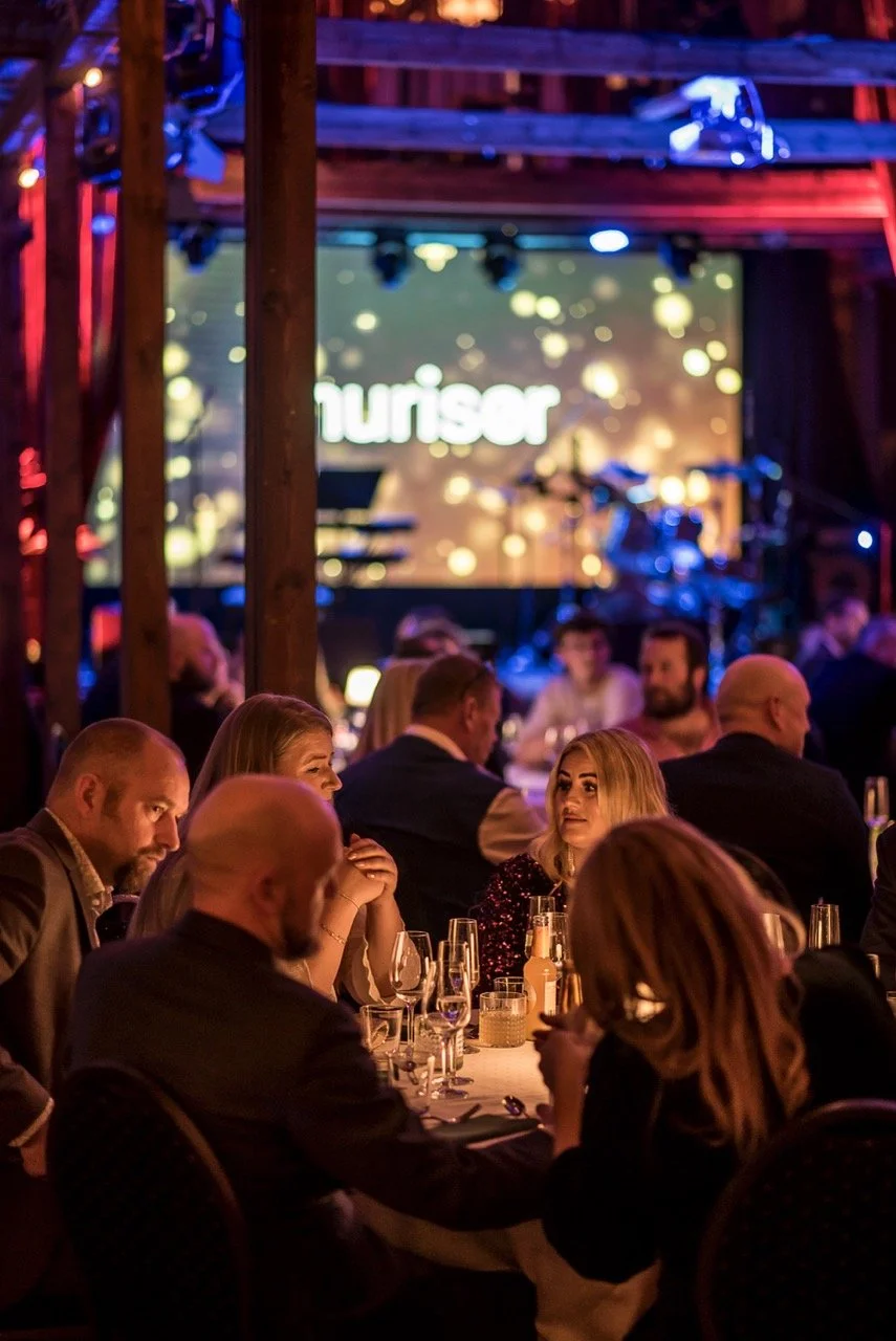 Mennesker som sitter rundt bord under et kveldsmåltid med musikkliv og en stor skjerm i bakgrunnen.