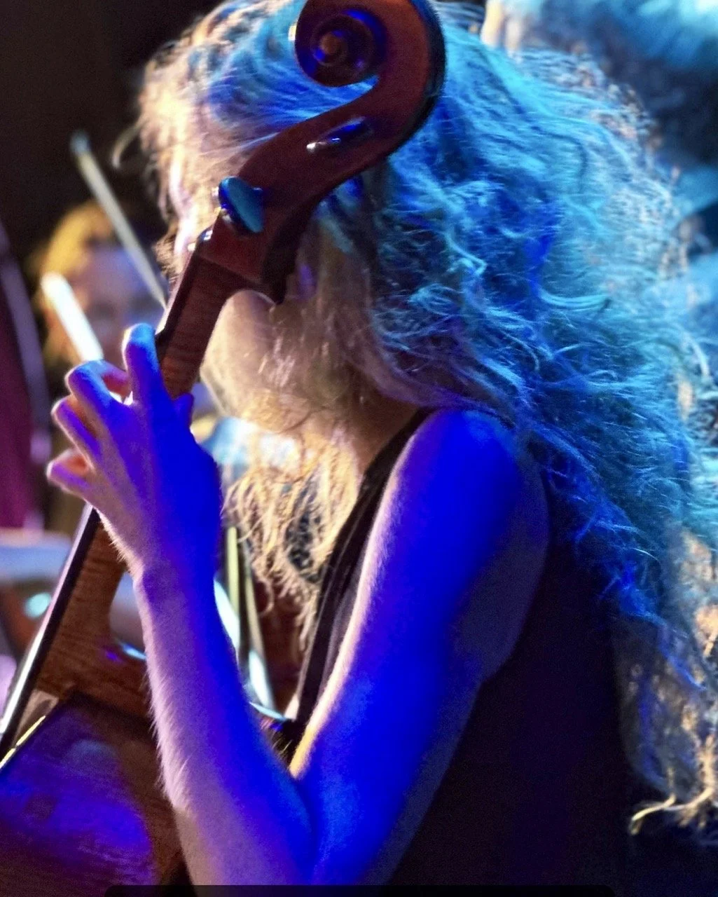 En person med lang, lyst hår spiller kontrabass i en mørk setting med blå belysning.