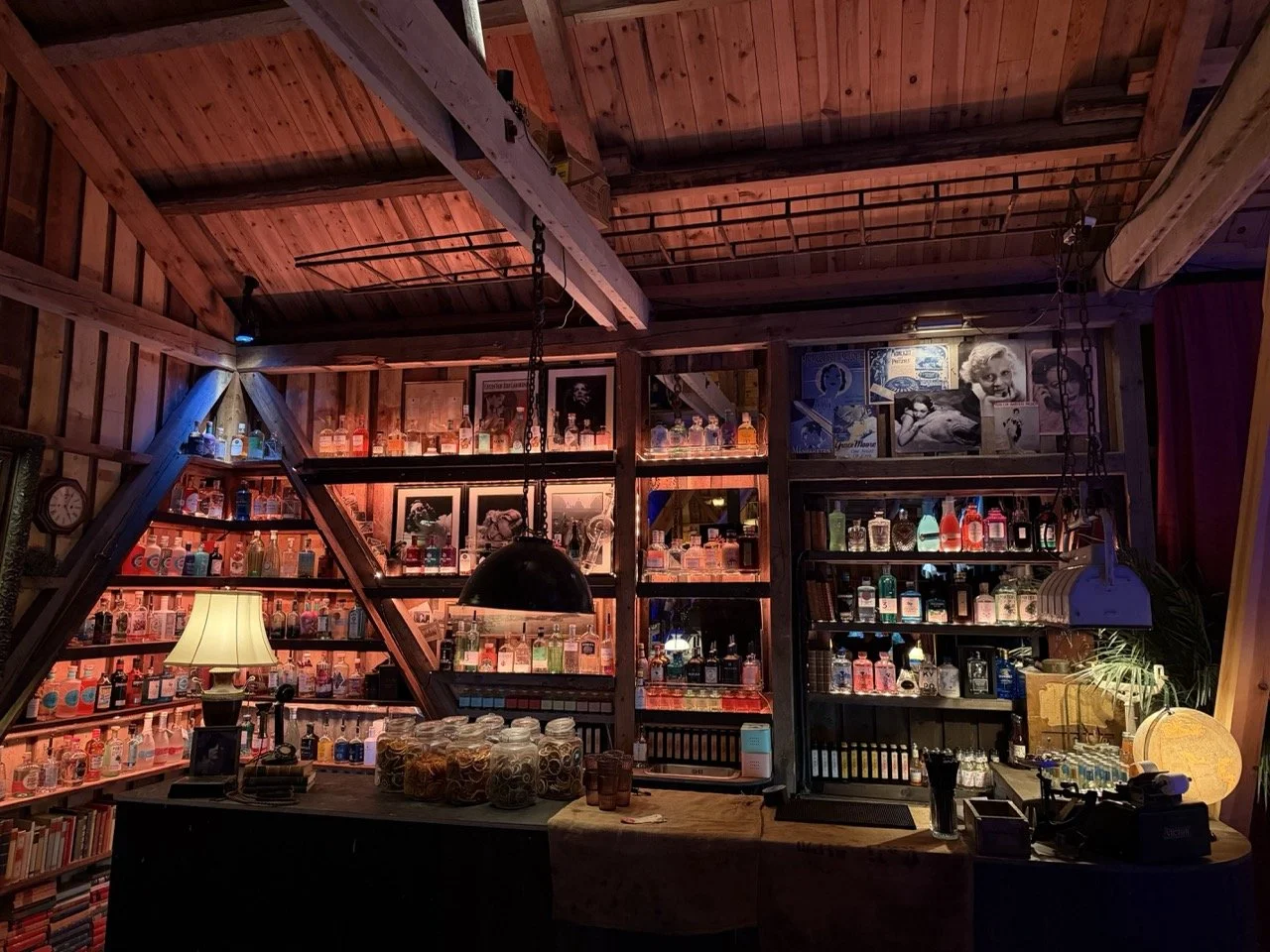 Innendørs barområde med trehyller fylt med forskjellige alkoholflasker, dekorative bilder på veggen, nattlampe, bord med kopper og diverse gjenstander.