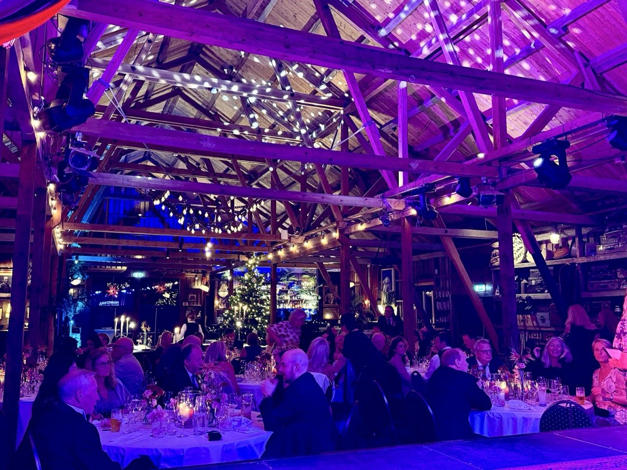 Innendørs julefest med mange mennesker, juletrær med lys, dekorasjoner, og bord med mat og drikke under tre-rammet tak.