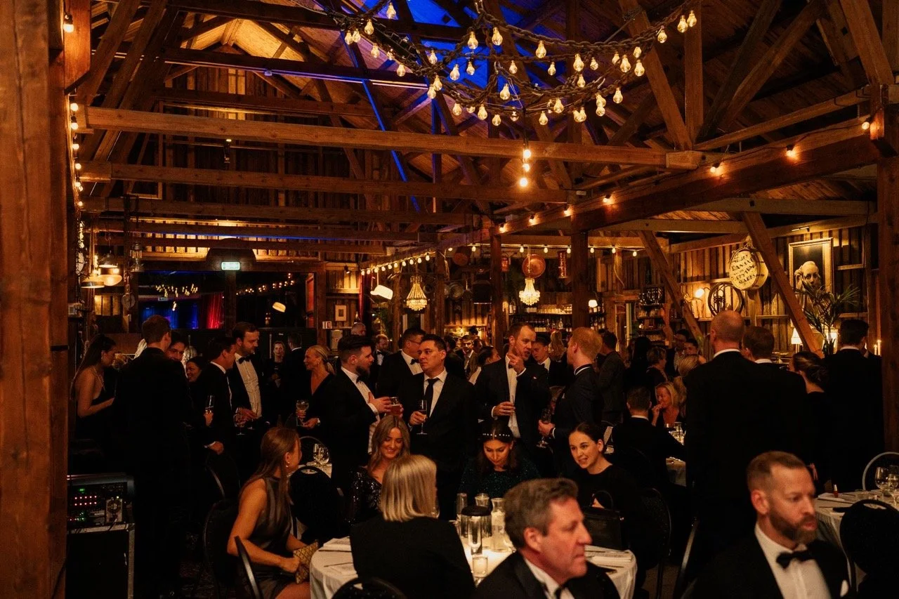 Mann og kvinner i formelle klær på en fest i en rustikk innendørs hall med trevegg, lysekrone, og dekorasjoner.