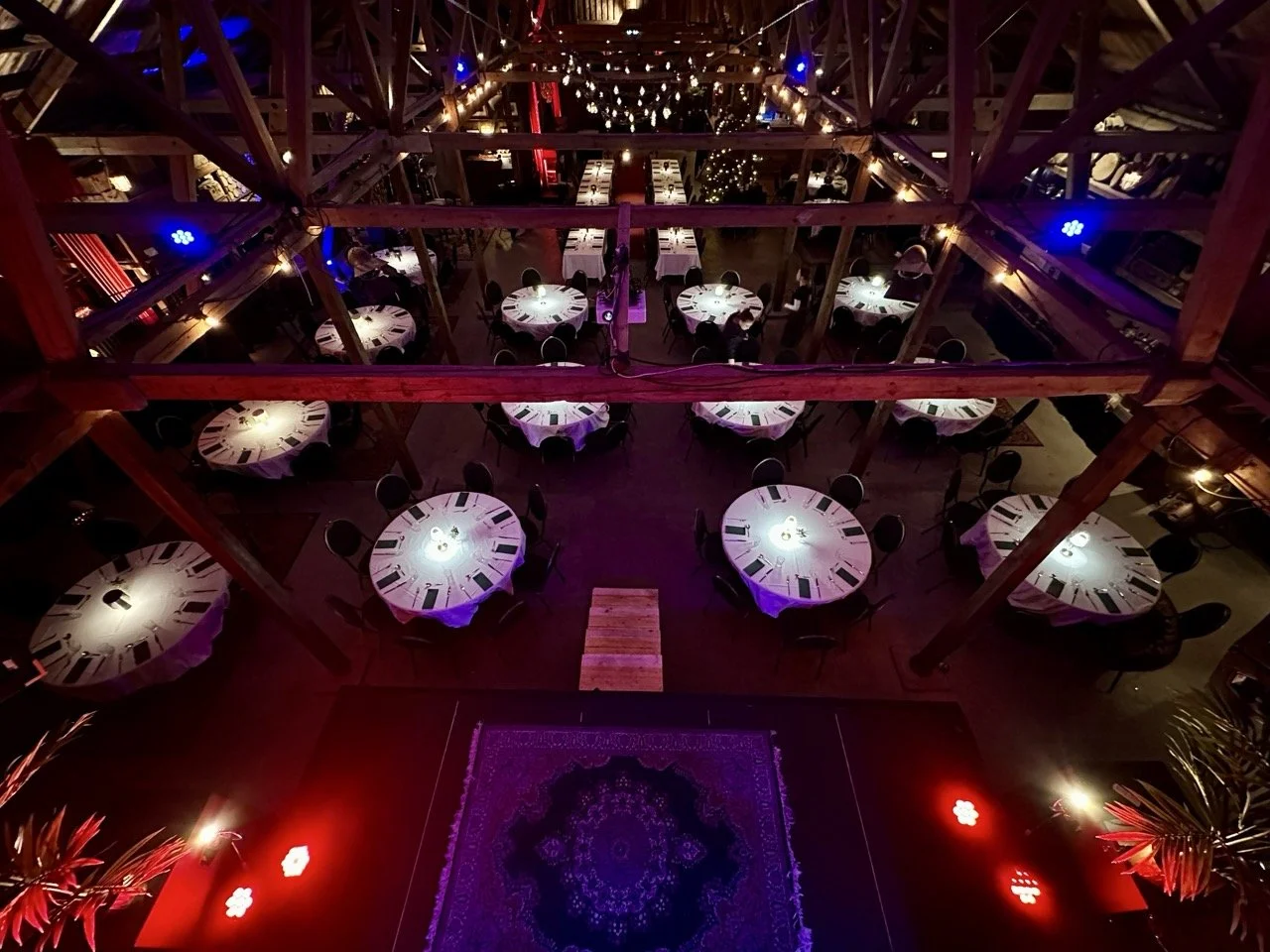 Innendørs selskapslokale sett fra en høyde med bord dekket til middag, dekorasjoner, belysning og et teppe foran en liten scene.