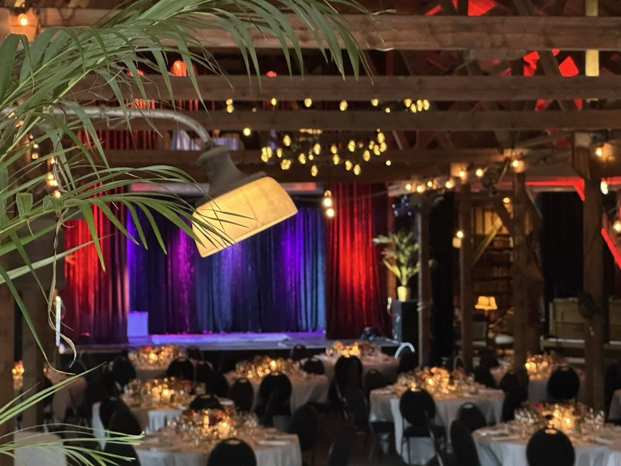 Innendørs festlokale med avrundede bord dekket med hvite duker, opplyst av små lyslykter. En scene med fargerike gardiner (blå, lilla og rød) og dekorativ belysning henger fra takbjelker. Det er bord med satengkjoler, og det er en stor plante i venstre side av bildet.