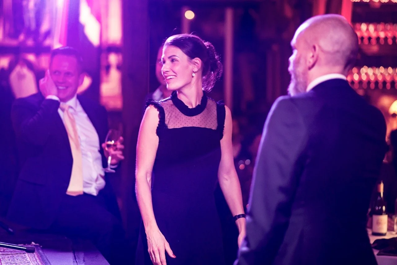 Tre personer på en fest, to menn og én kvinne, i formelle klær, smiler og snakker i et rom med rosa og lilla belysning.