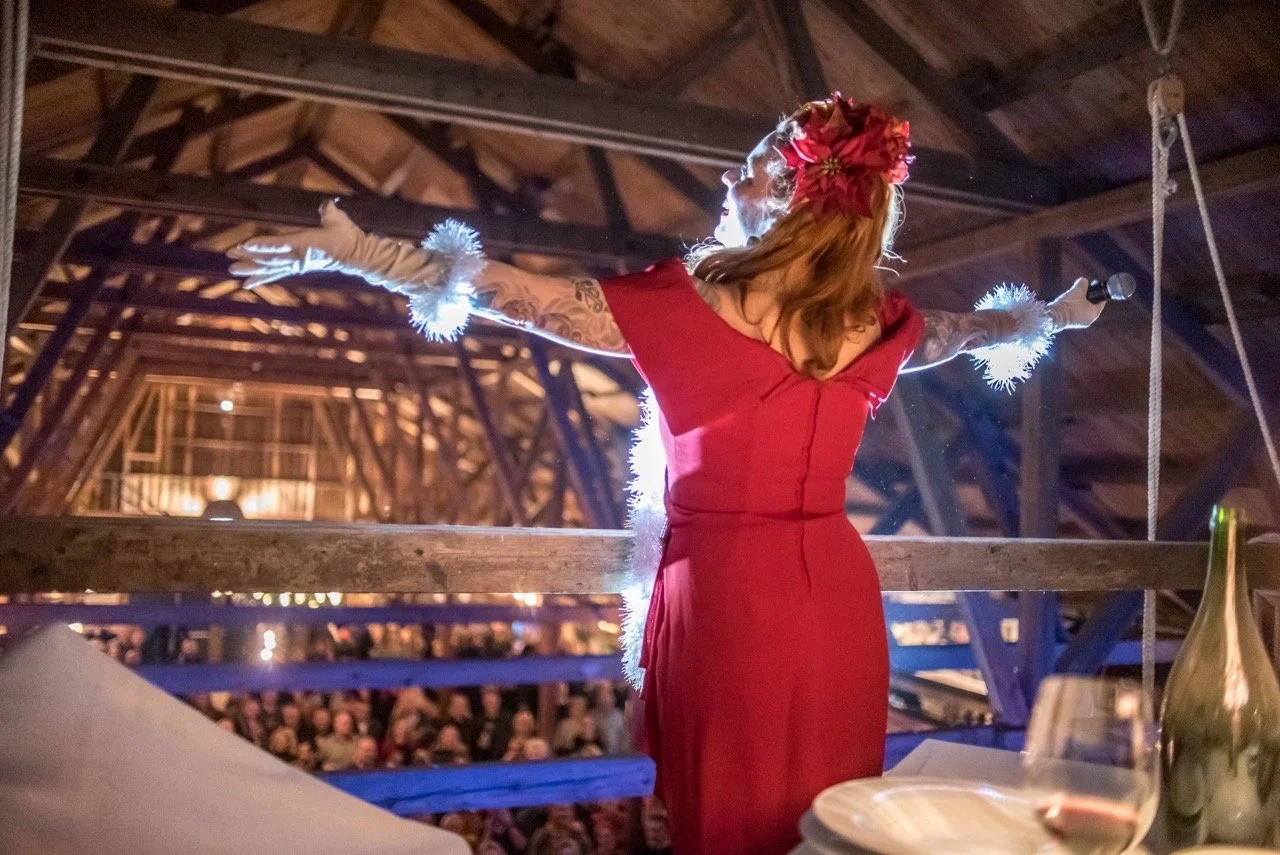 En kvinne i en rød kjole med blomster i håret, holder en mikrofon og står med armene utstrakt foran en stor forsamling i et treverkstak, sannsynligvis under en arrangement.