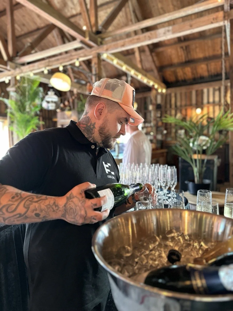 En mann med tatoveringer og en brenton caps heller opp champagne i et glass i en rustikk innredning med grønne planter.
