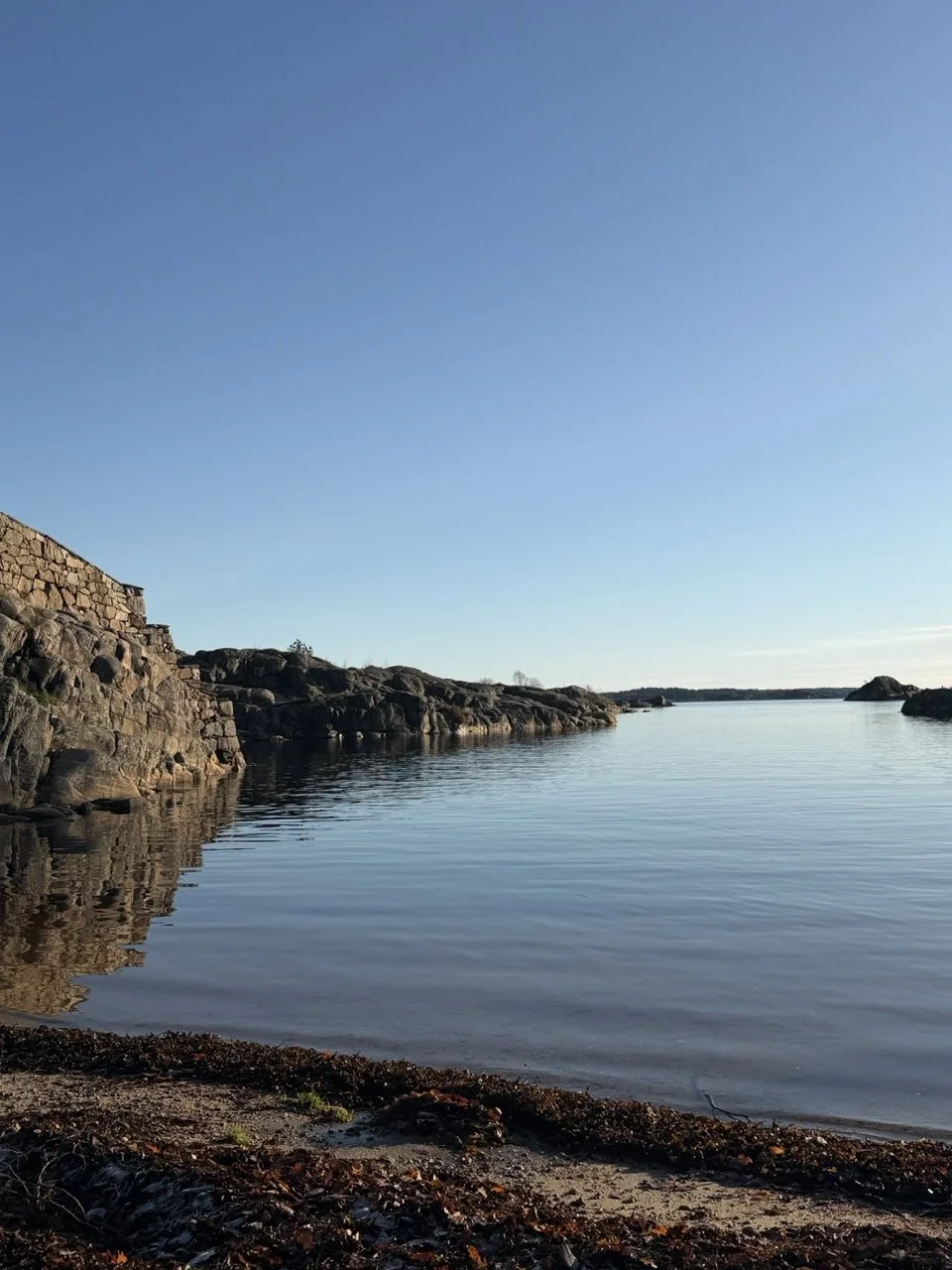 Bilde av en rolig sjø med stein- og klippeformasjoner langs stranden under en klar blå himmel.