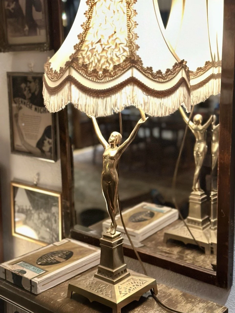 En dekorativ bordlampe med en gullfarget kvinnefigur som holder en lampskjerme med blonder og pynt, plassert foran en speil og et bord med bøker.