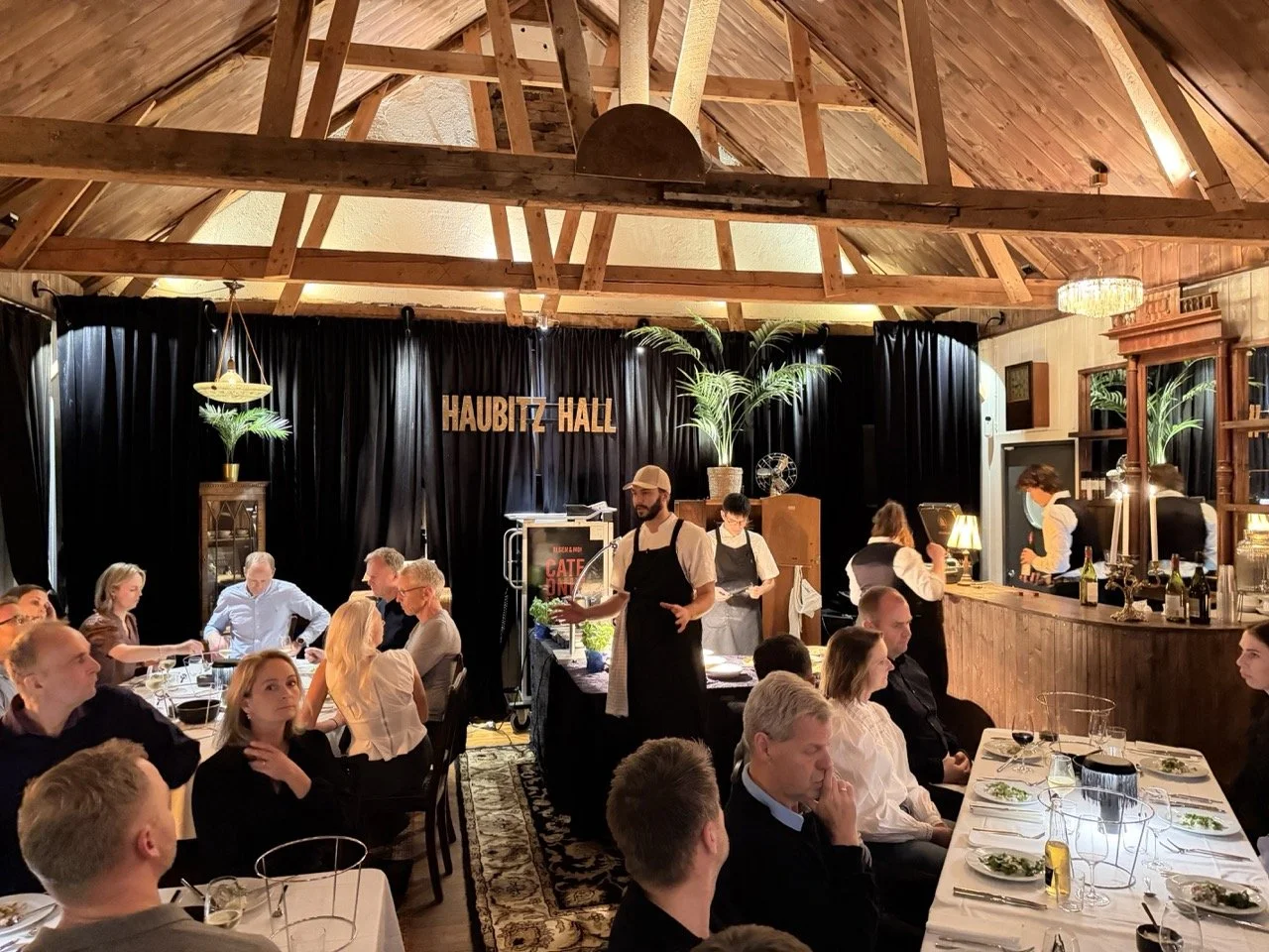 Interiør av Haubert Hall med gjester som spiser middag, et dekorativt innredningsområde med planter, og en servitør som serverer mat.