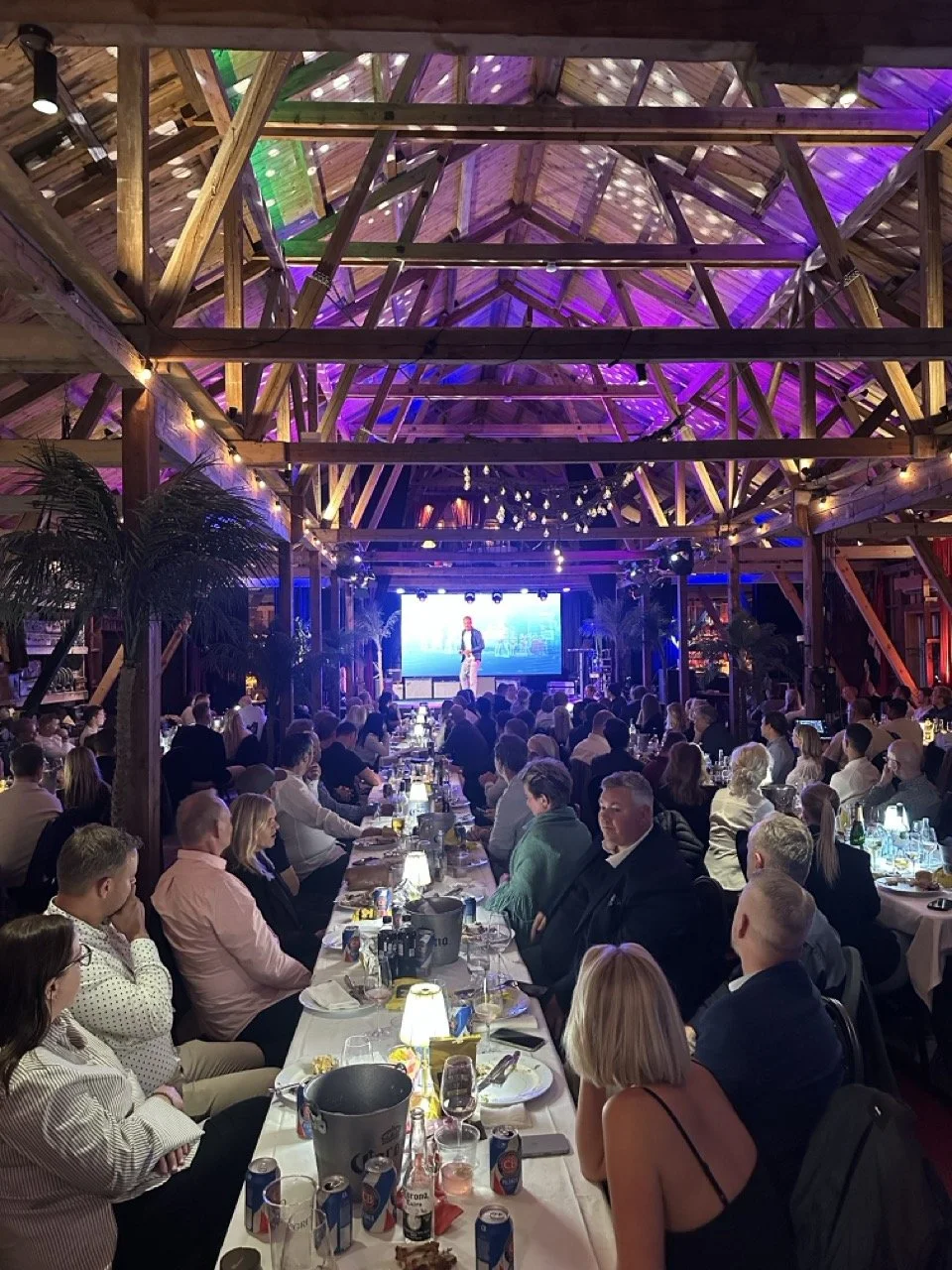 Innendørs scene av en stor gruppe mennesker som sitter ved langbord, ser på en scene med en person som holder en presentasjon. Det er treverk i taket og stemningen er festlig med fargerik belysning og dekorasjoner.