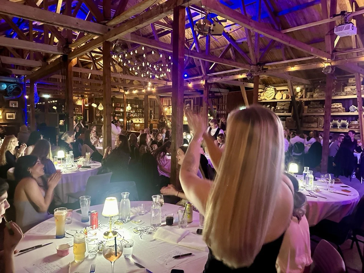 Middagsselskap i et rustikt trehus med mange mennesker som ler og har det gøy, dekorert med lyslenker, borddekorasjoner og opplysningsutstyr.