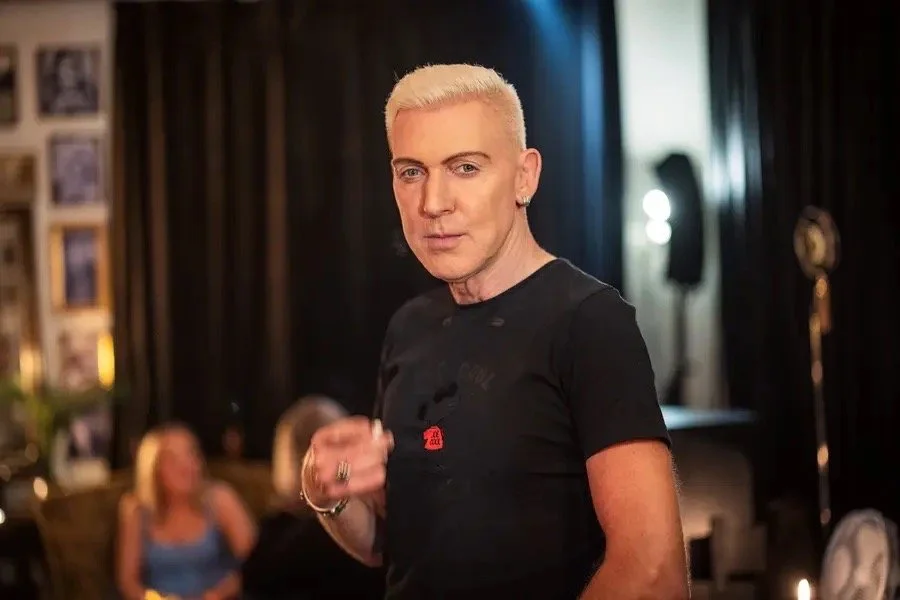 En person med bleket hår og svart T-skjorte står i en romslig, mørklagt lokal med folk i bakgrunnen.