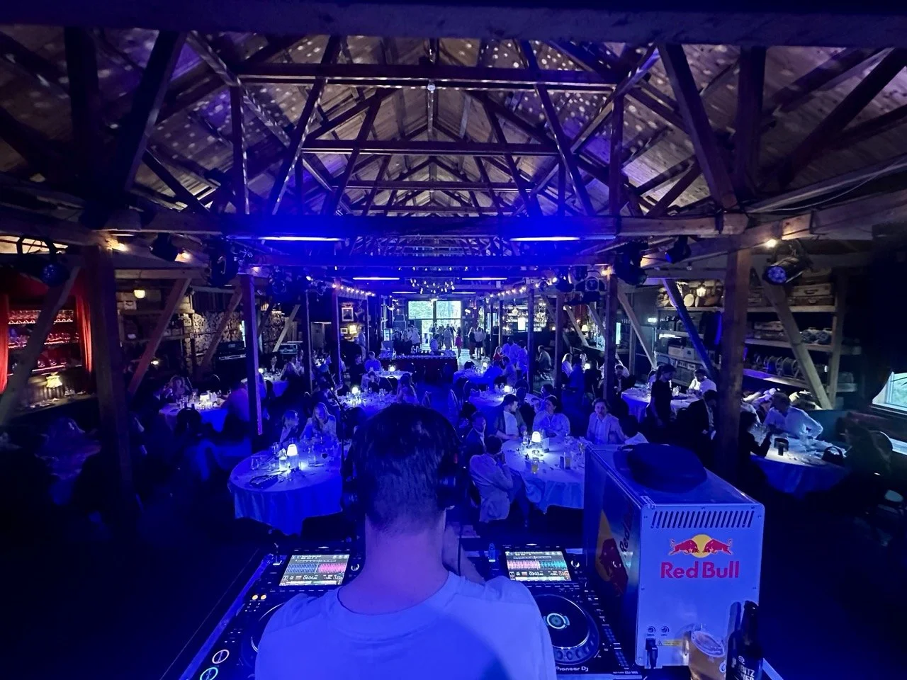 Innendørs event i en tømret hytte med flere gjester som sitter ved byrundebord. DJ står foran og spiller musikk i et opplyst rom med blå og lilla lys.