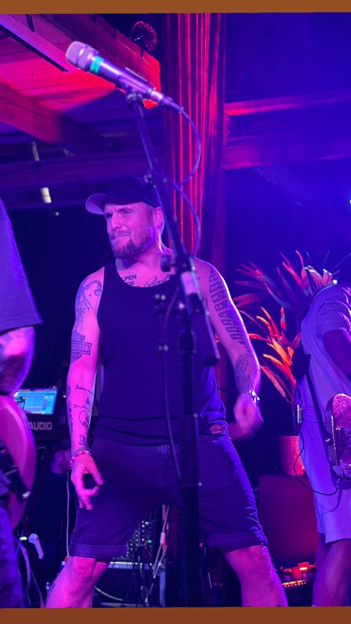 En mann med tatoveringer, mørke klær og caps, som opptrer på en scene med mikrofon foran seg, under farget belysning.