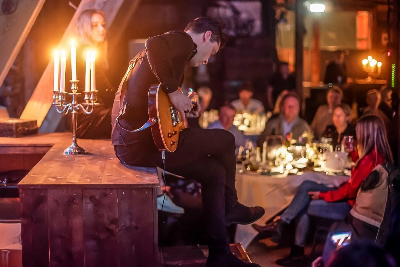 En mann spiller elektrisk gitar på en scene med et bord med lyslenker foran og publikum som ser på i et rustikt, koselig lokale.