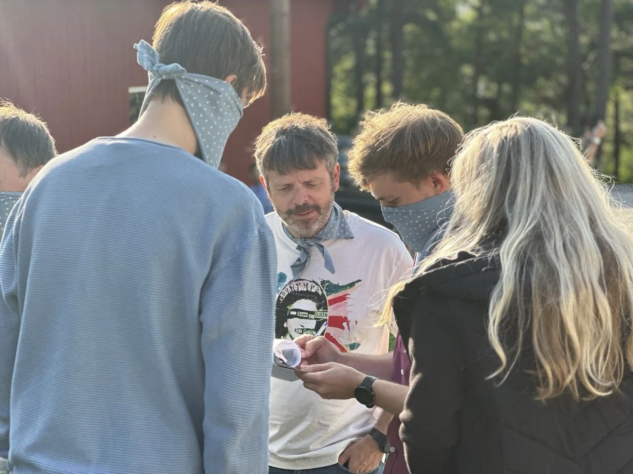 Gruppe mennesker, fire personer iført grå ansiktsmasker, ser på noe på en telefon utendørs med trær i bakgrunnen