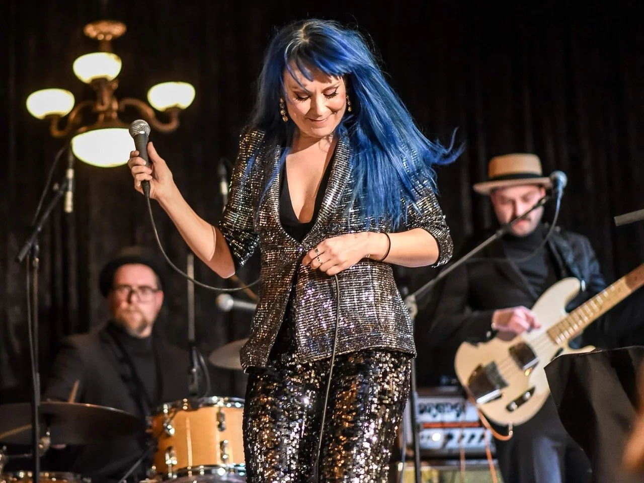 En kvinne med blå hår som synger eller opptrer på scenen, med to menn som spiller instrumenter i bakgrunnen.