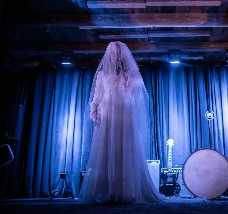 En person kledd i brudekjole og brudeblondet slør står på en scene med blå belysning, omgitt av musikkutstyr og instrumenter.