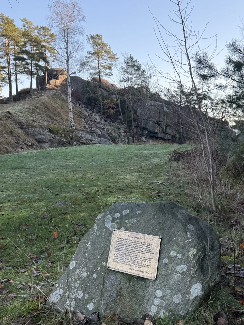 En stor stein med en informasjonstavle på foran, omgitt av gress og spredte blader. Bak er det en skråning med trær og fjell i bakgrunnen, med noen løvtrær uten blader og en forseggjort bygning eller struktur på toppen.