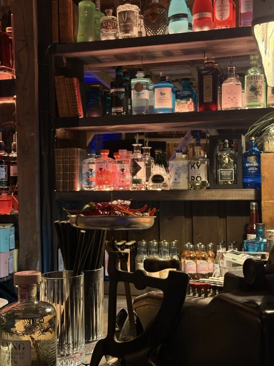 Innendørs bar med hyller fullt av ulike spritflasker, lave glass med rosa drikke, og barutstyr.