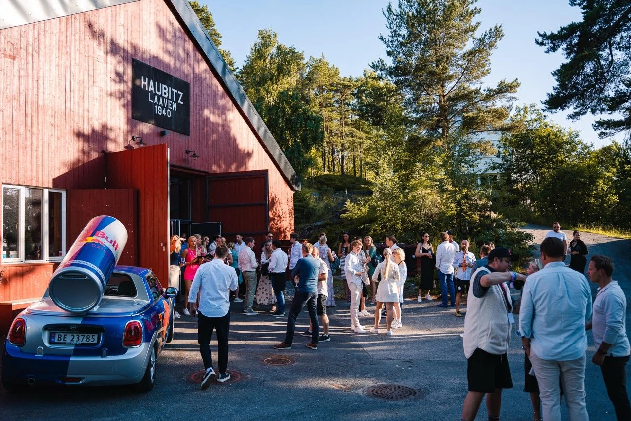 Mannsamling utenfor en rød bygning med et stort Red Bull-ølkan på en bil foran. Gjestene er kledd i formelle klær, og det er solskinn med grønne trær i bakgrunnen.
