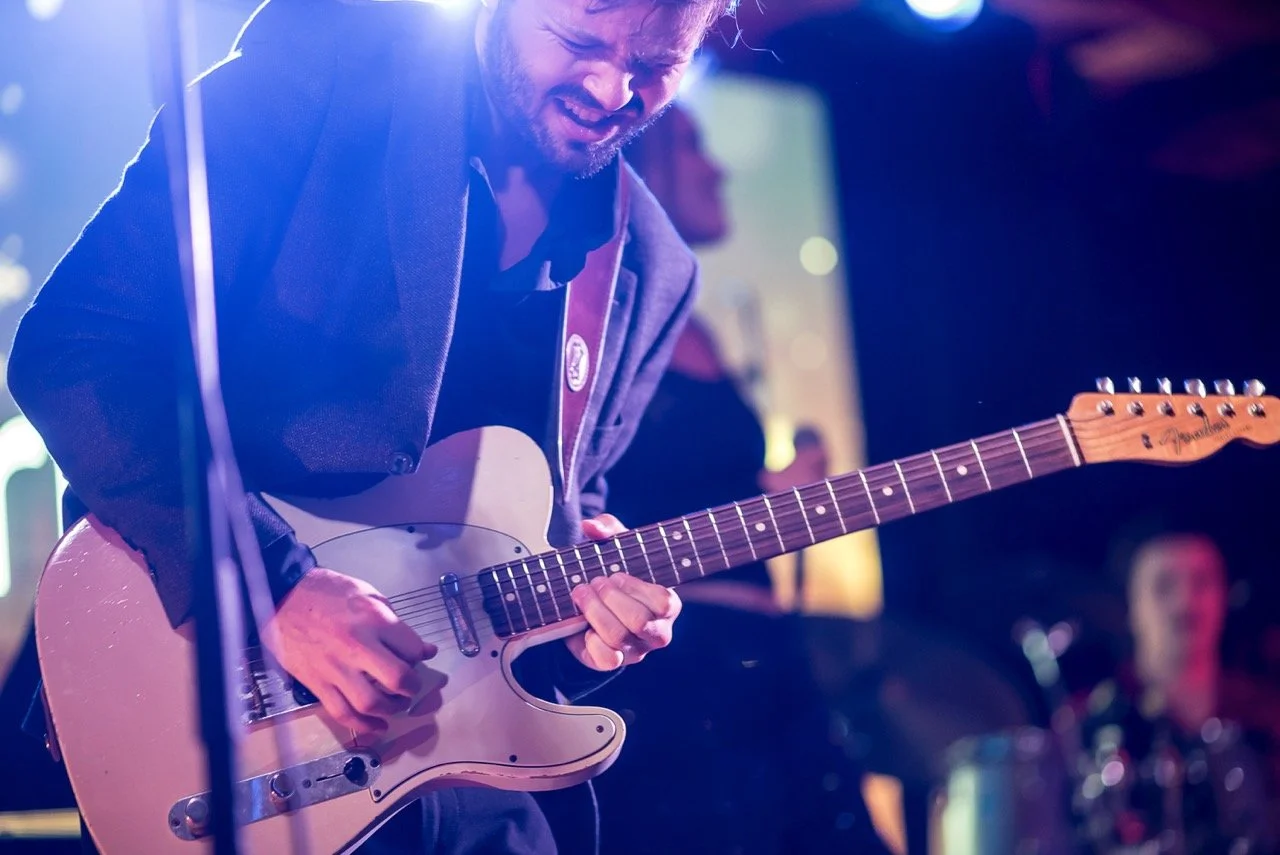 En mann spiller på en elektrisk gitar under en konsert, med fokuserte uttrykk og fargerike lys i bakgrunnen.