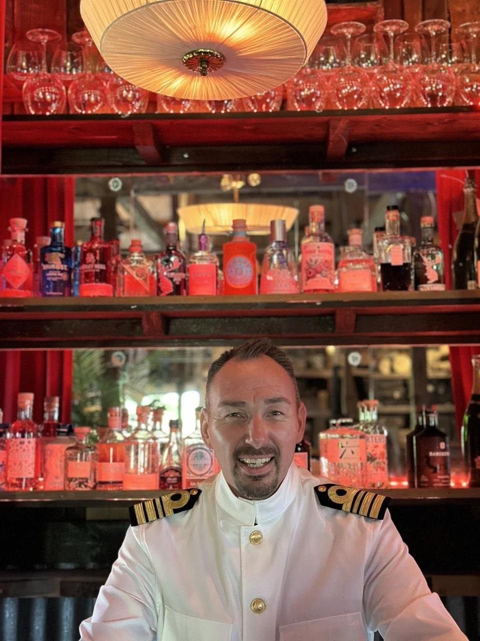 En mann kledd i en hvit uniform med gullknapper og skulderepauleter sitter foran en bakke med ulike flasker med røde etiketter, på en bar. Over ham henger det en taklampe og glass på en hylle.