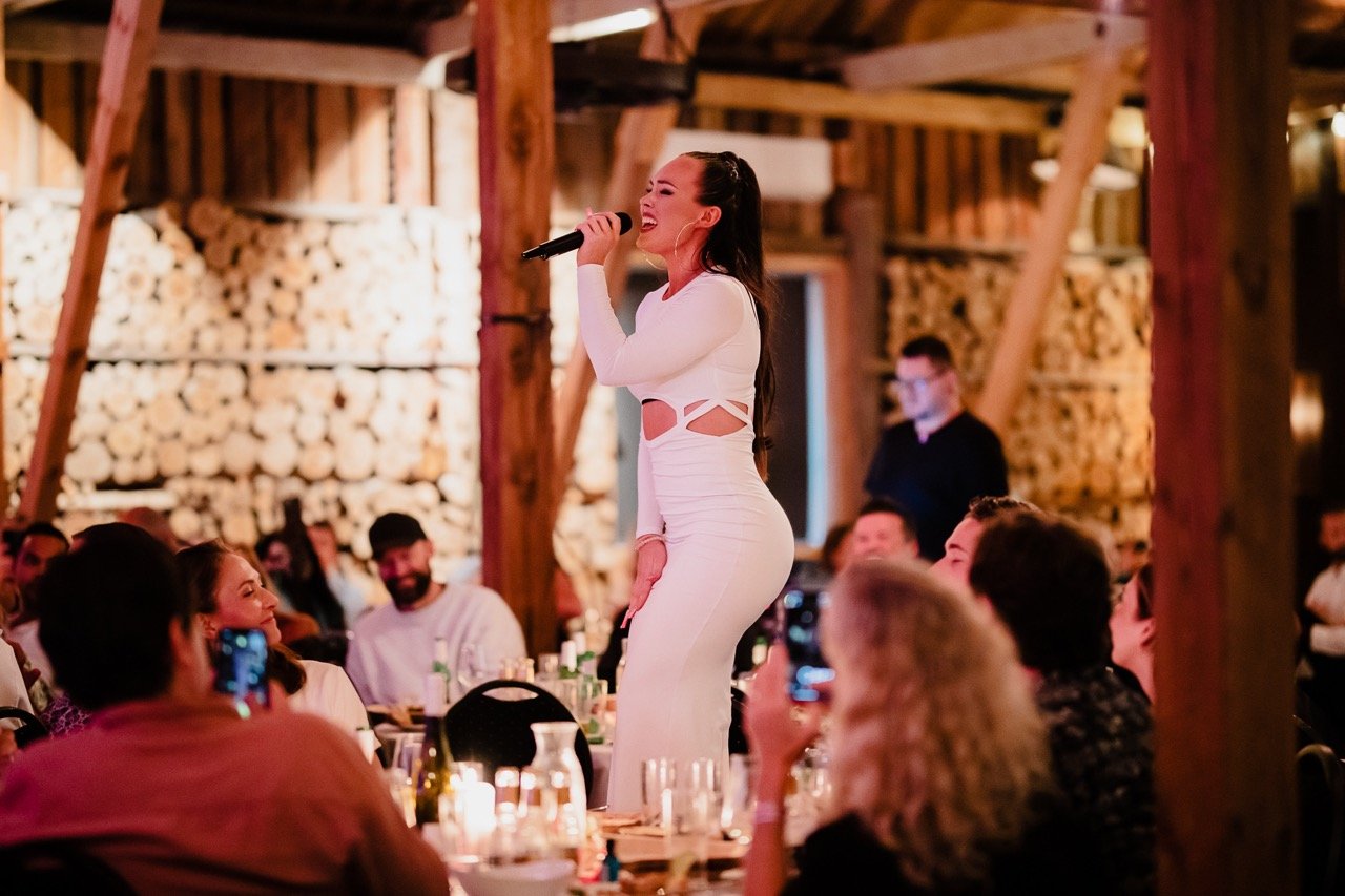 En kvinne i en hvit festkjole synger med mikrofon mens hun står på et bord foran en mengde mennesker i et rustikt rom med trekonstruksjoner og fyringsved i bakgrunnen.