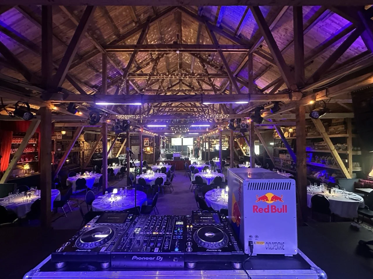 Interiør av et rustikk konsert- eller eventlokale med tre takbjelker, belysning i blå og lilla nyanser, og runde bord med hvit duker og tent lys. DJ-utstyr i forgrunnen og en bar med flasker i bakgrunnen.
