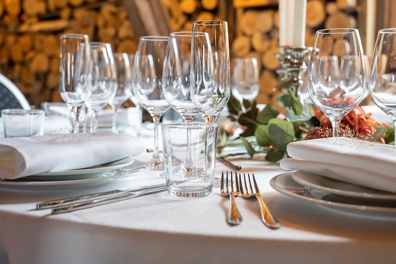 Elegant middagsbord dekket med hvit duk, flere tomme vinglass, vann glass, sølvbestikk og hvite tallerkener med serviett, dekorert med grønne blader og blomster i bakgrunnen.