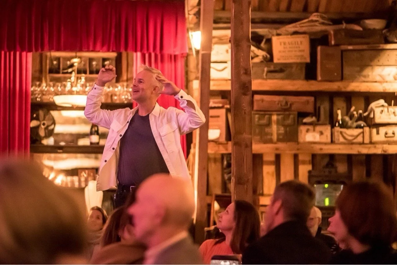 En mann står foran en gruppe mennesker i en innendørs setting, visst nok en bar eller en restaurant, med røde gardiner og hyller med ulike gjenstander i bakgrunnen. Han smiler og har hendene opp i luften, muligens midt i en tale eller en forestilling