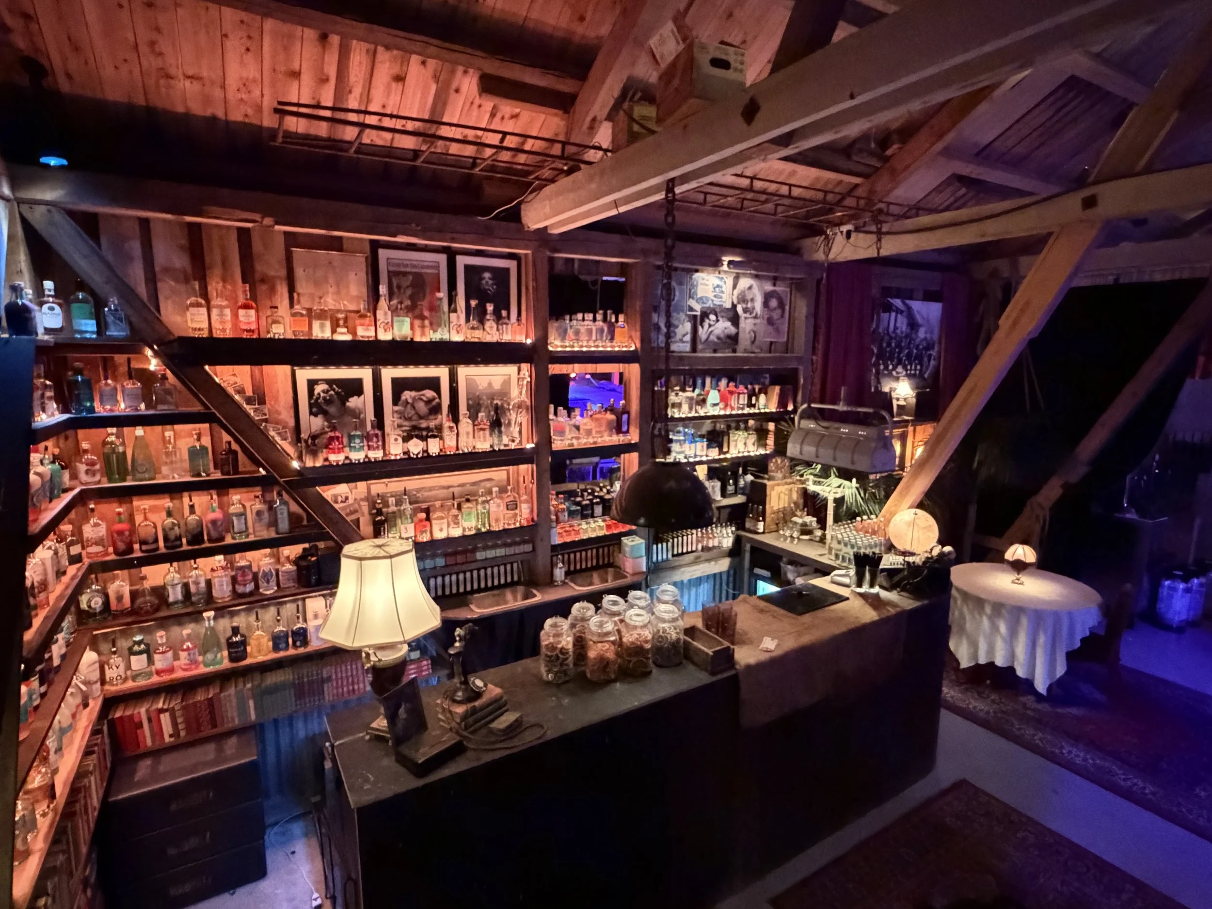 Et koselig hjemmekino med bar med ulike alkoholflasker på hyllene, lamper og bilder på veggene, og et bord dekket med et duk. Rødt, blått og oransje belysning skaper en stemningsfull atmosfære.