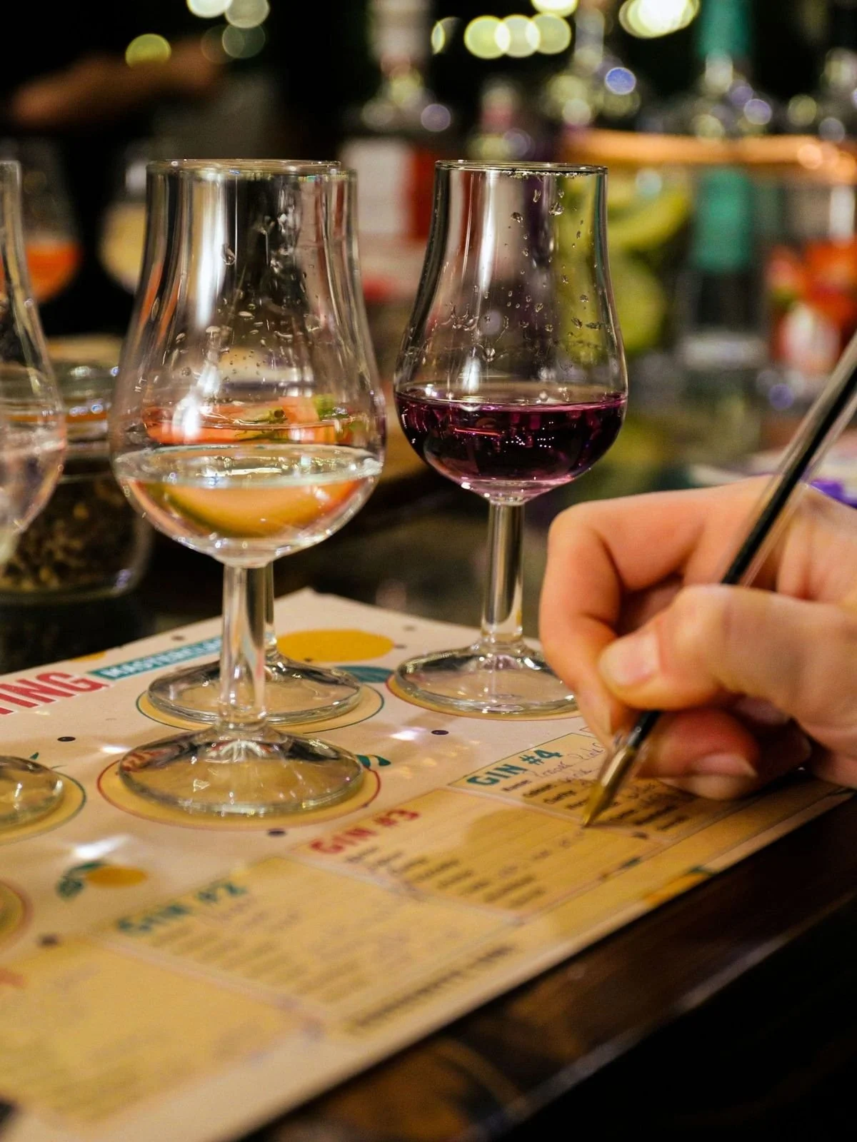 To vinglass med klar væske og ett vinglass med rød vin. En person skriver på en smaksskjema på en bar eller restaurant.