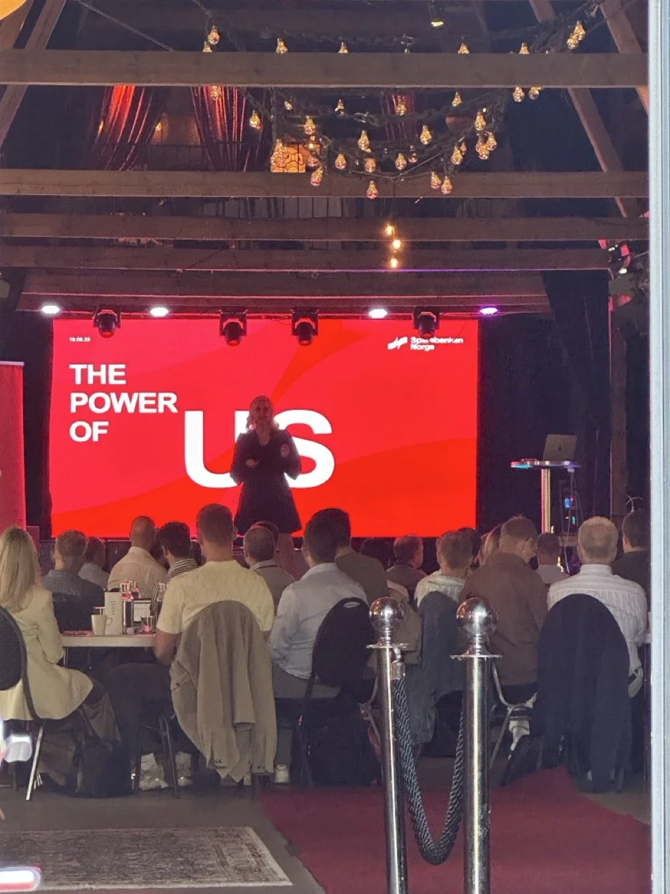 En gruppe mennesker deltar på en konferanse med en presentasjon på storskjerm som sier 'The Power of US'.