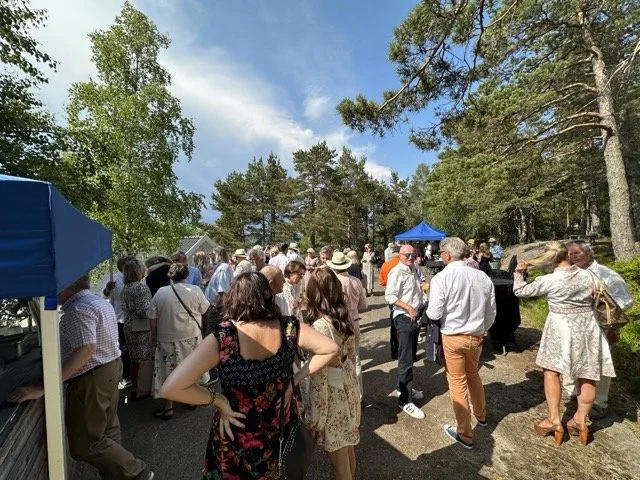 Grupp mennesker samlet utendørs, noen står og snakker, det er en blå parasoll og trær i bakgrunnen.