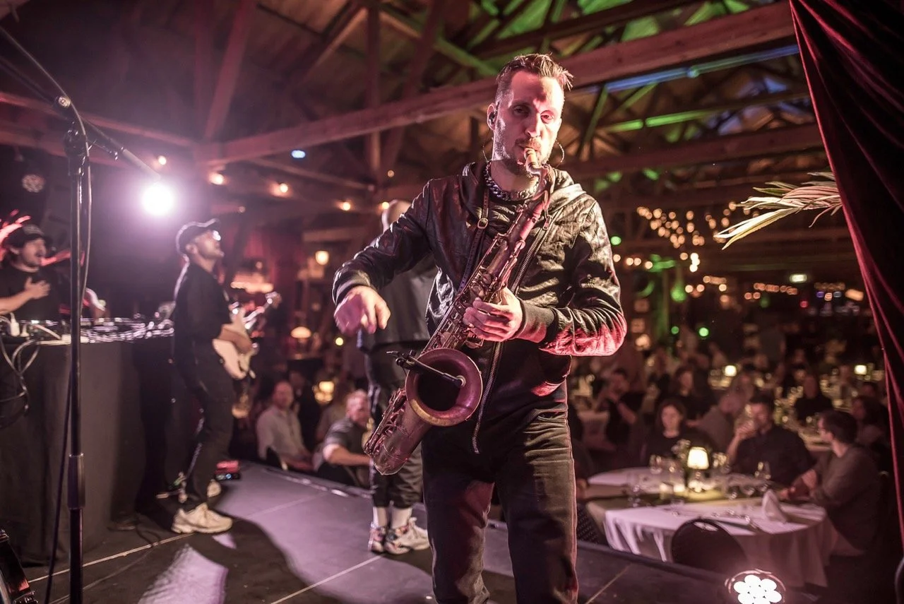 En mann som spiller saksofon på scenen i en restaurant eller bar, med et publikum i bakgrunnen og rommet dekorert med belysning og planter.