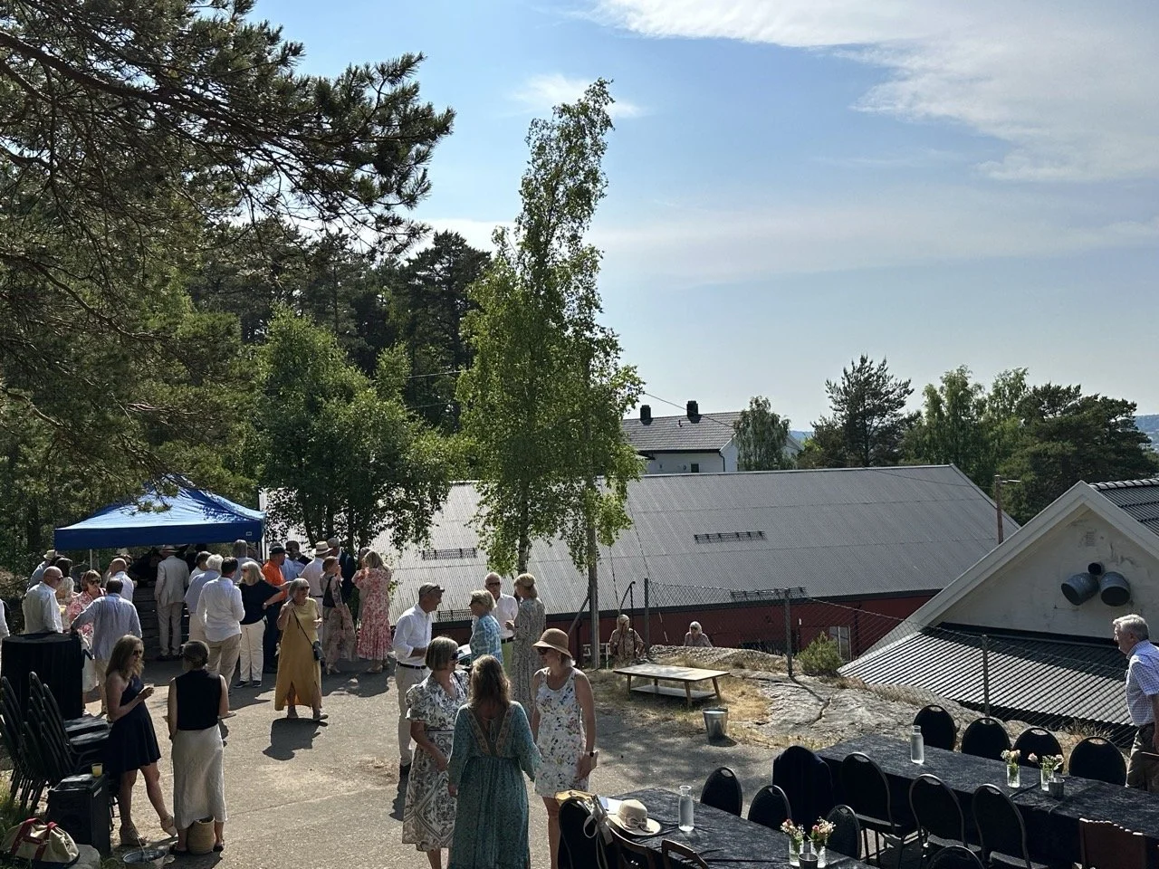 En gruppe mennesker samles utendørs under en solemn eller sosial begivenhet, med noen rundt et bord og andre under en blå telt. Det er trær og bygninger i bakgrunnen, og værforholdene er solfylte.
