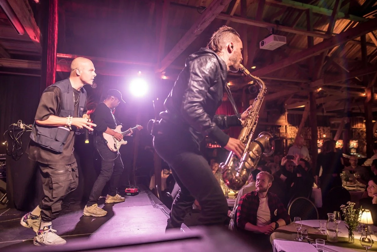 Musikere spiller opp på en scene i et rustikt lokale med treverk og myk belysning, og publikum ser på.