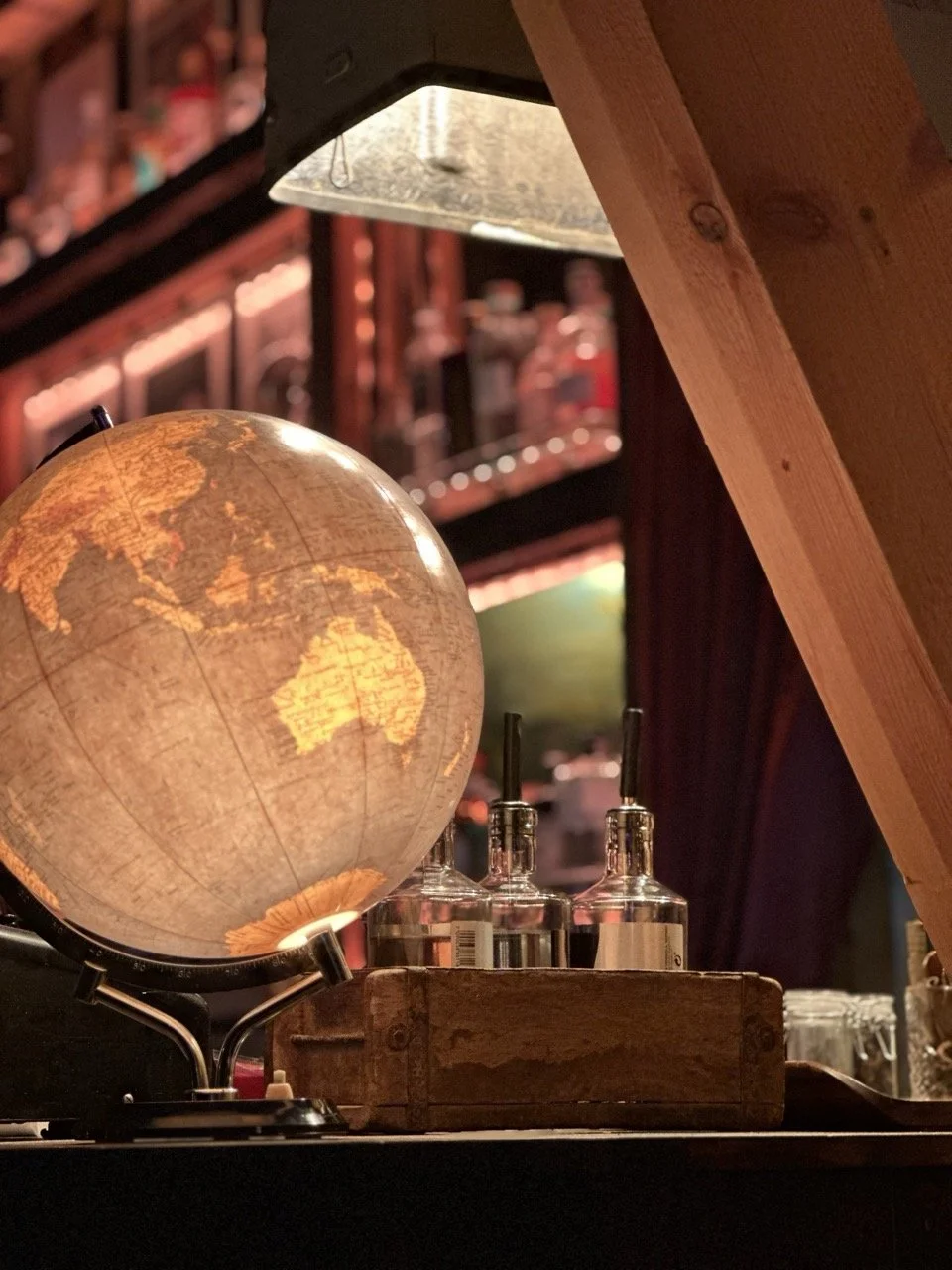 En bord med en opplyst verden globe, to sylindriske glassflasker med metalltut, en trekasse og andre barartikler under en skrånende takbjelke i en bar eller kafé.