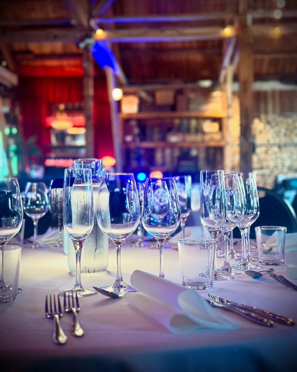 Bord dekket for et selskap med tomme vinglass, bestikk og serviett, på en uteservering med trestak og dekorative lys i bakgrunnen.