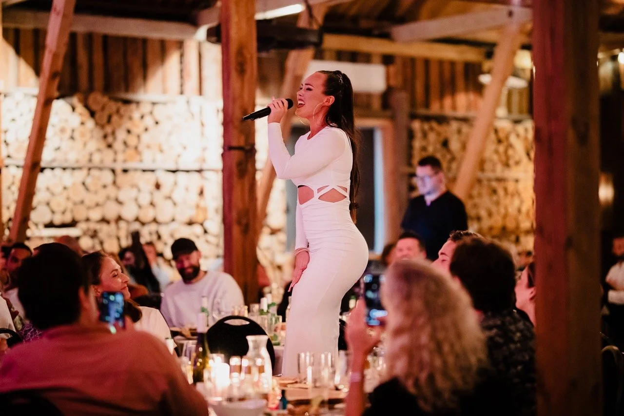 En kvinne synger med mikrofon foran et publikum i en rustikk innendørs setting, omgitt av mennesker som tar bilder og ler.