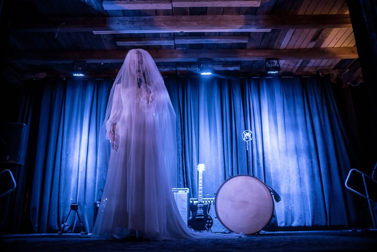 En scene med et hvitt brudekjole, gitar, tromme og mikrofon foran blå gardiner, under teppe av tre.